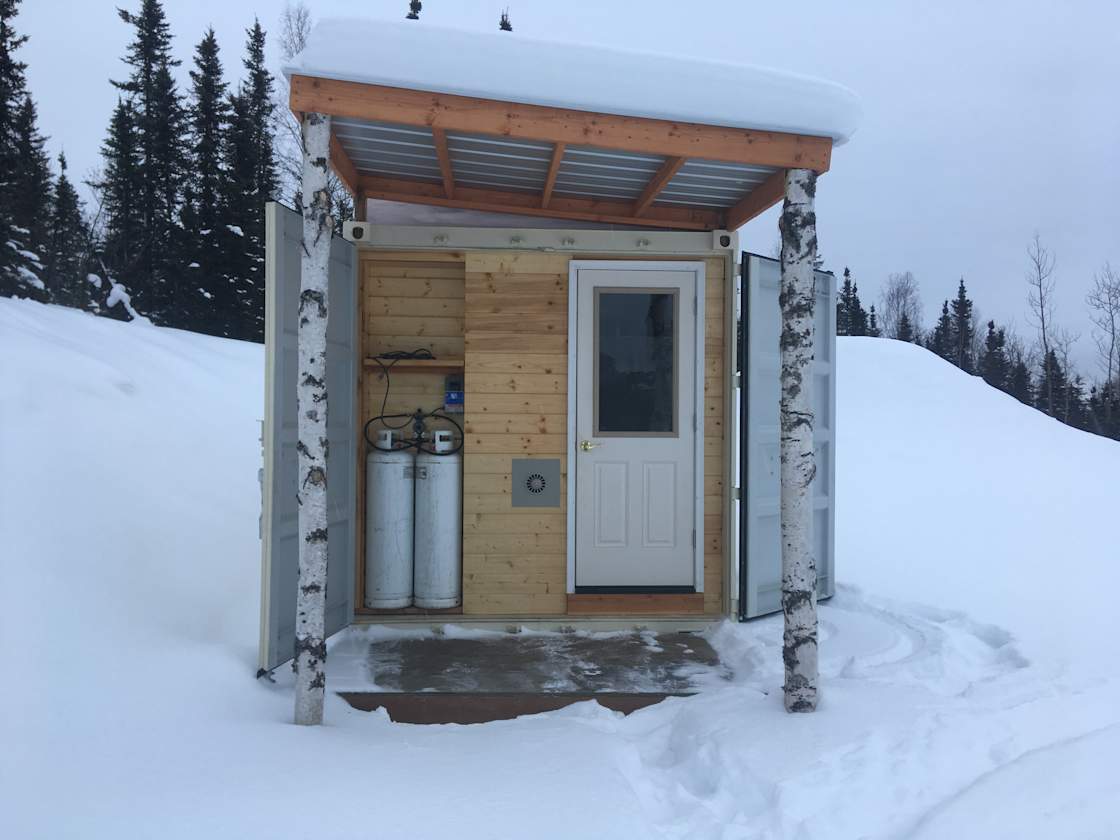 This tiny house is a converted shipping container. It has a small covered porch, a locking front door and two propane tanks that fuel the kitchen range, heater, and solitary light.