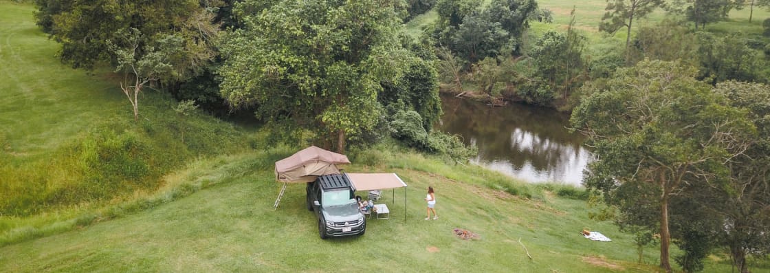 Aerial view of a peaceful riverside camping spot with a 4x4, roof tent, awning, and a picnic blanket, surrounded by lush greenery and trees