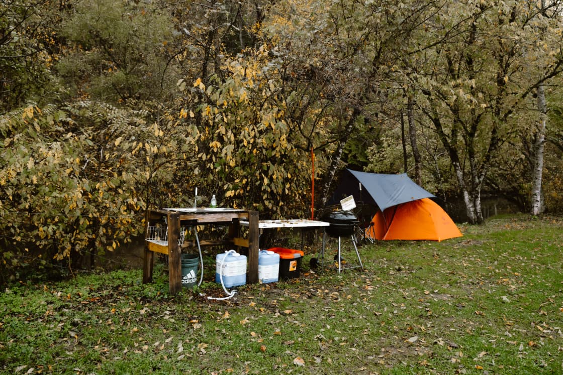 The campsite includes a spot for your tent, a fire pit, a hammock hanger, a picnic table, and a pump-sink next to a charcoal grill.