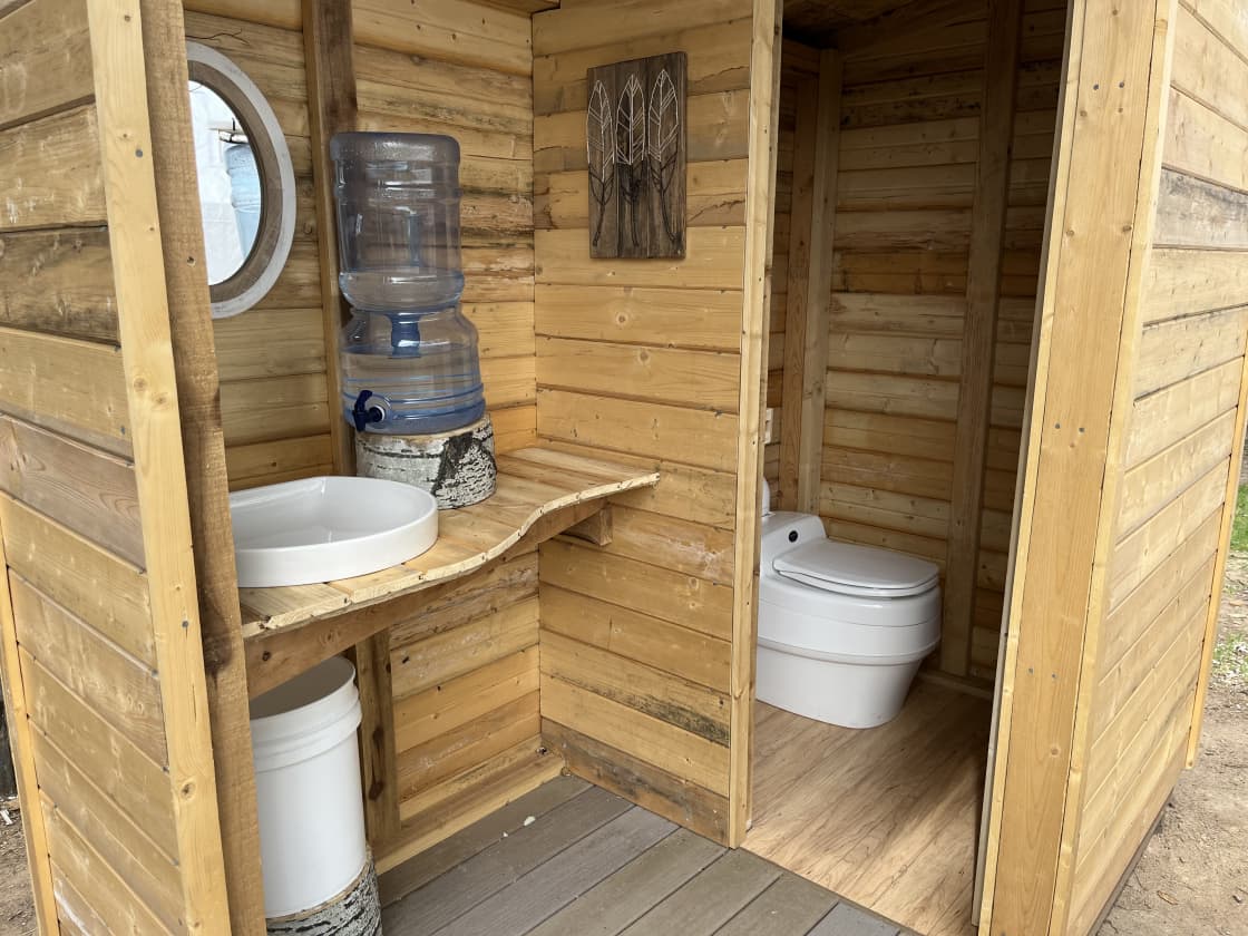 Outhouse with countertop, sink, mirror, water jug, and composting toilet.