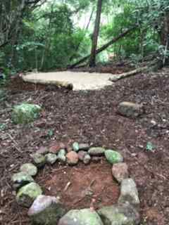 Cleared camp site surrounded by forest, prepared fire pit area close by.