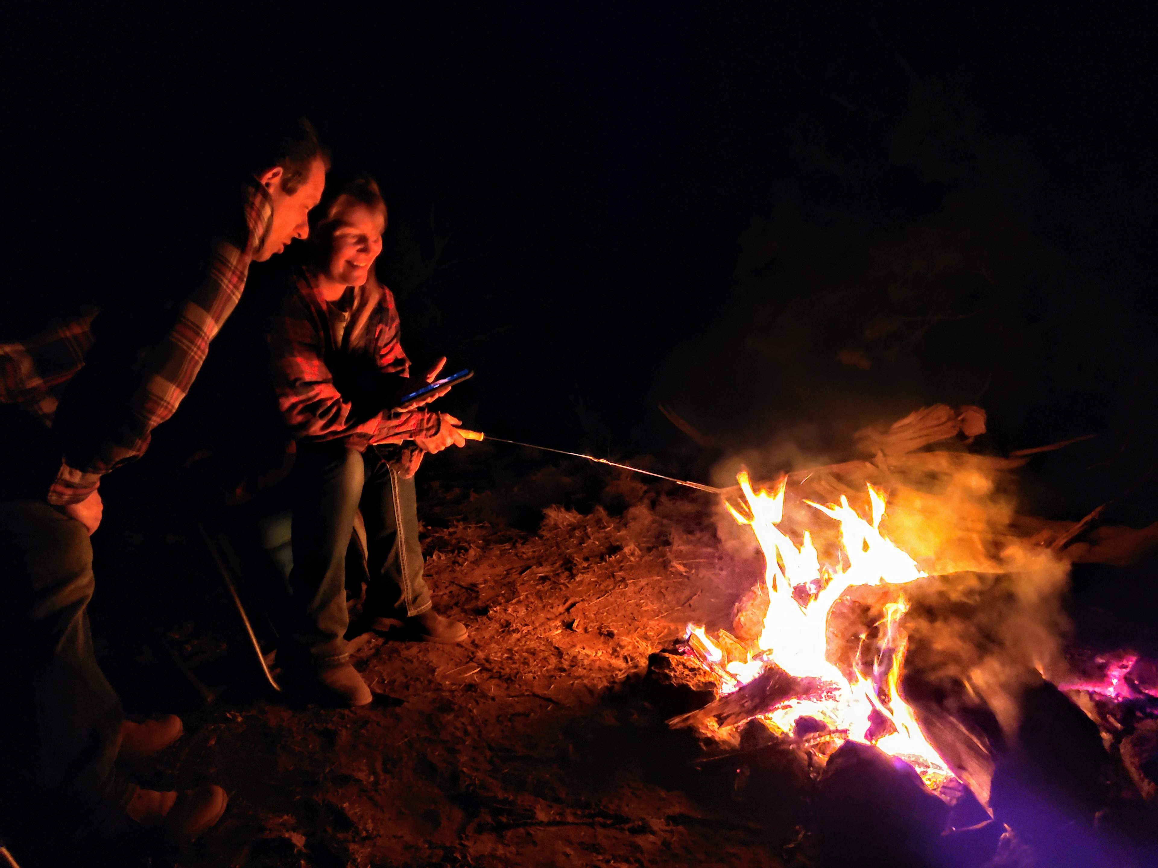 A couple of campers roasting marshmallows and video-chatting with their family by the fire.