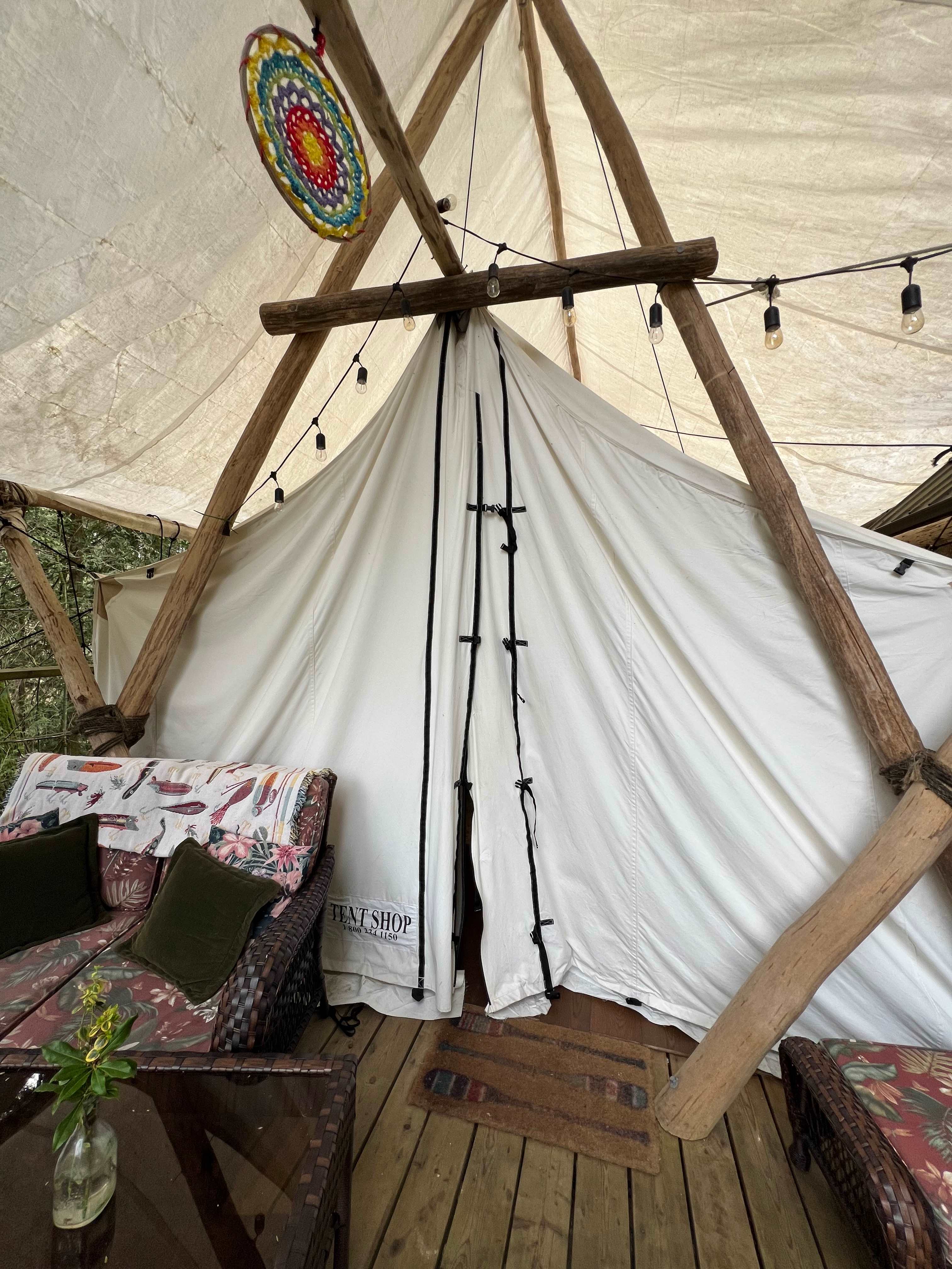 A white canvas tent pitched with bleached, smooth lumber, under a similar canopy. String lights hang decoratively above the tent wings, and a rainbow dreamcatcher hangs above the centre. A wicker loveseat set is placed in front of the tent, with matching table