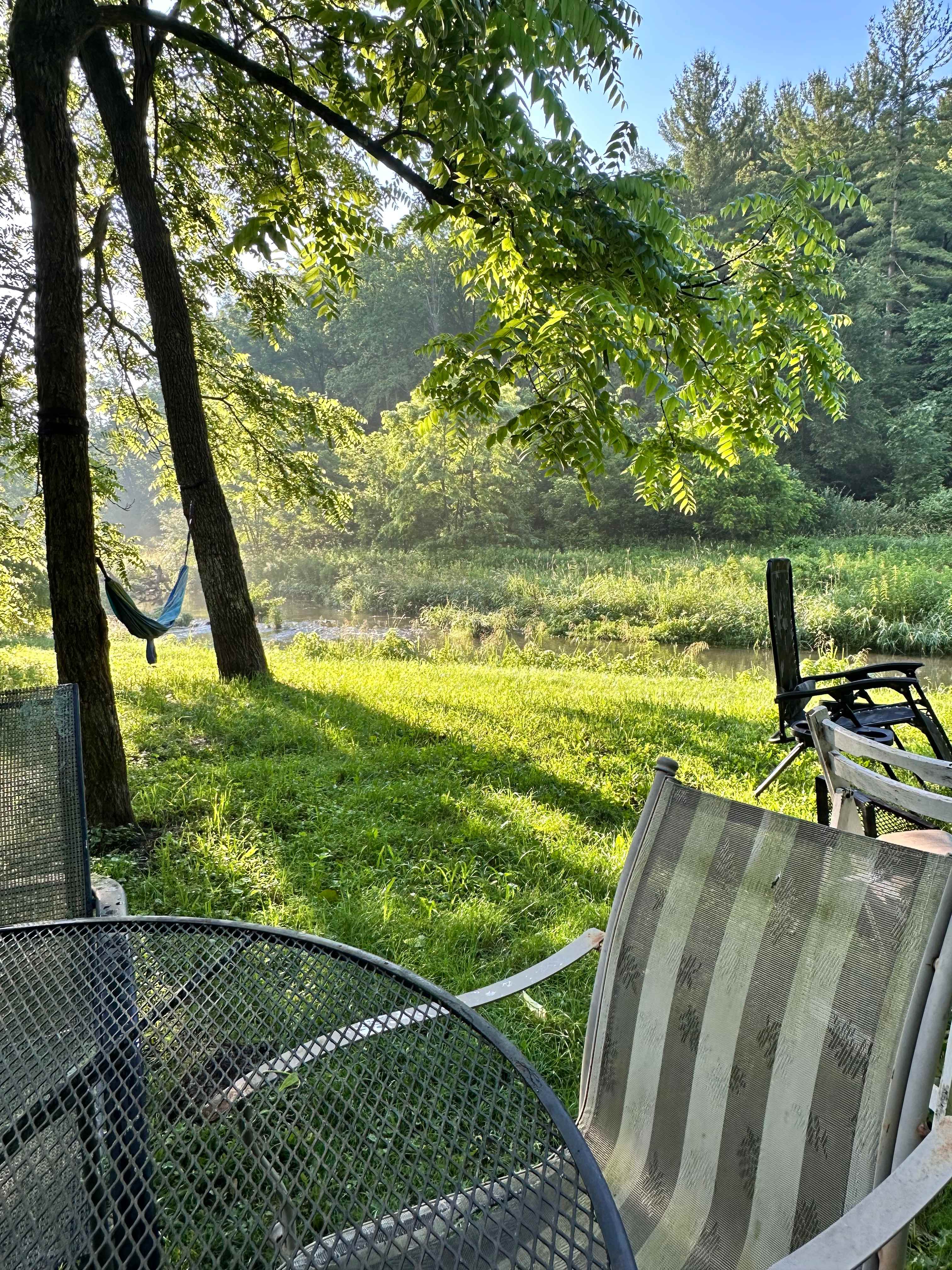 Patio furniture, hammock, and chair around fire ring visible facing the stream.