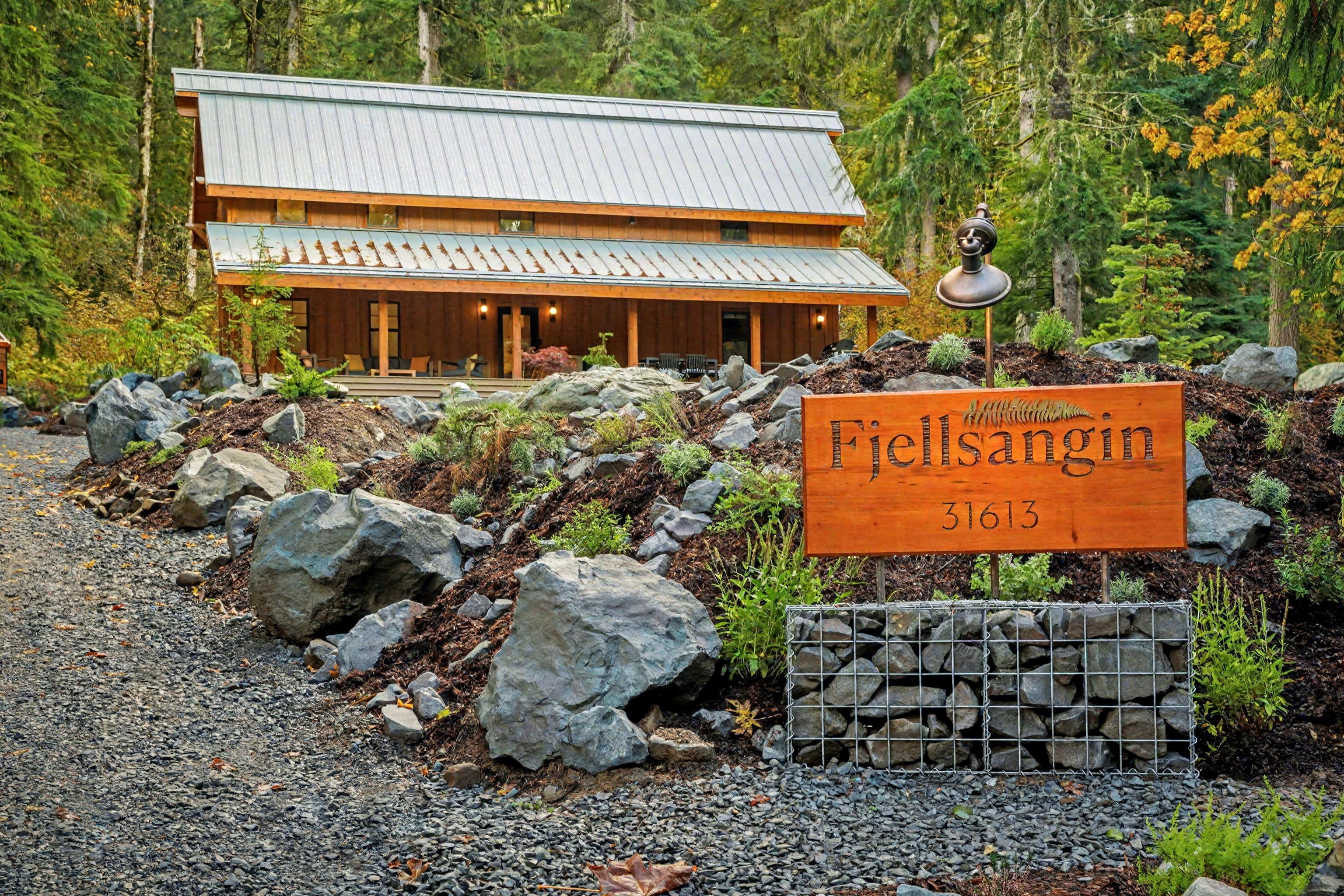 The exterior of Fjellsangin nestled into the forest, with the hand-crafted sign, gabion stone landscaping, and towering old-growth firs framing the ro