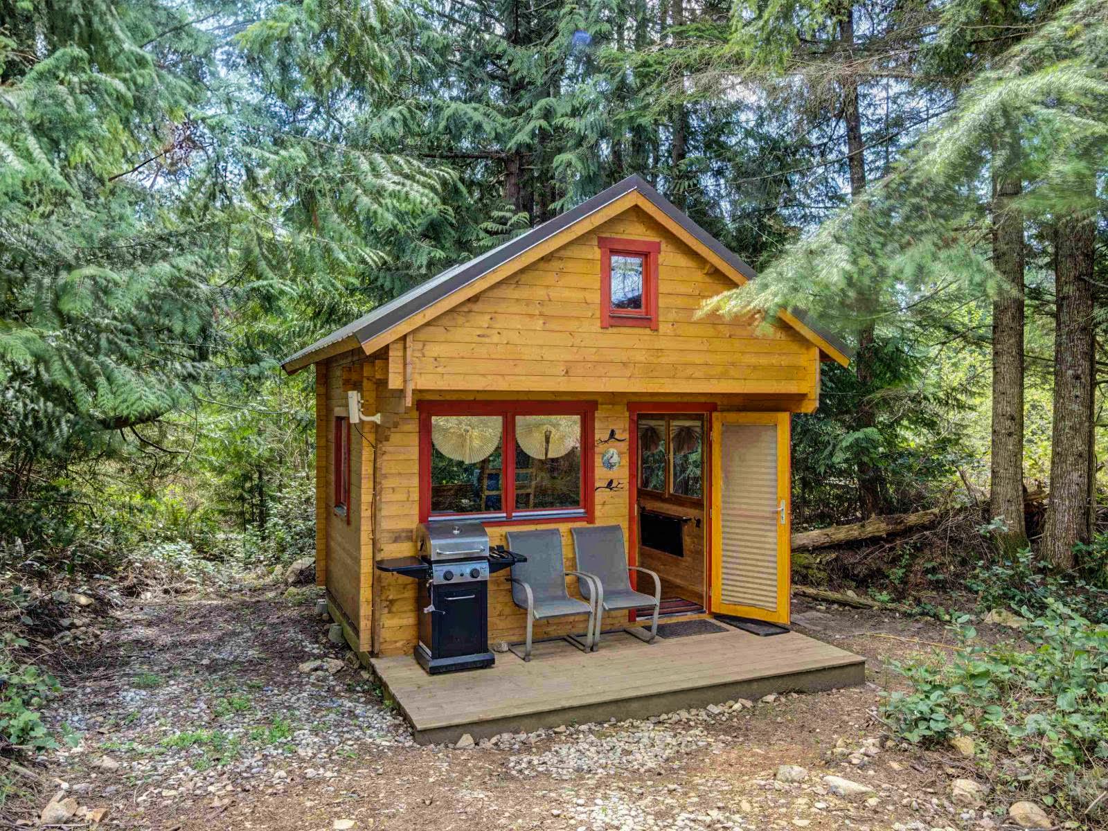 The Peacock cabin is nestled in the woods. The front deck features a barbecue and two comfy chairs. There is a separate area on the same site with an outdoor dining table.