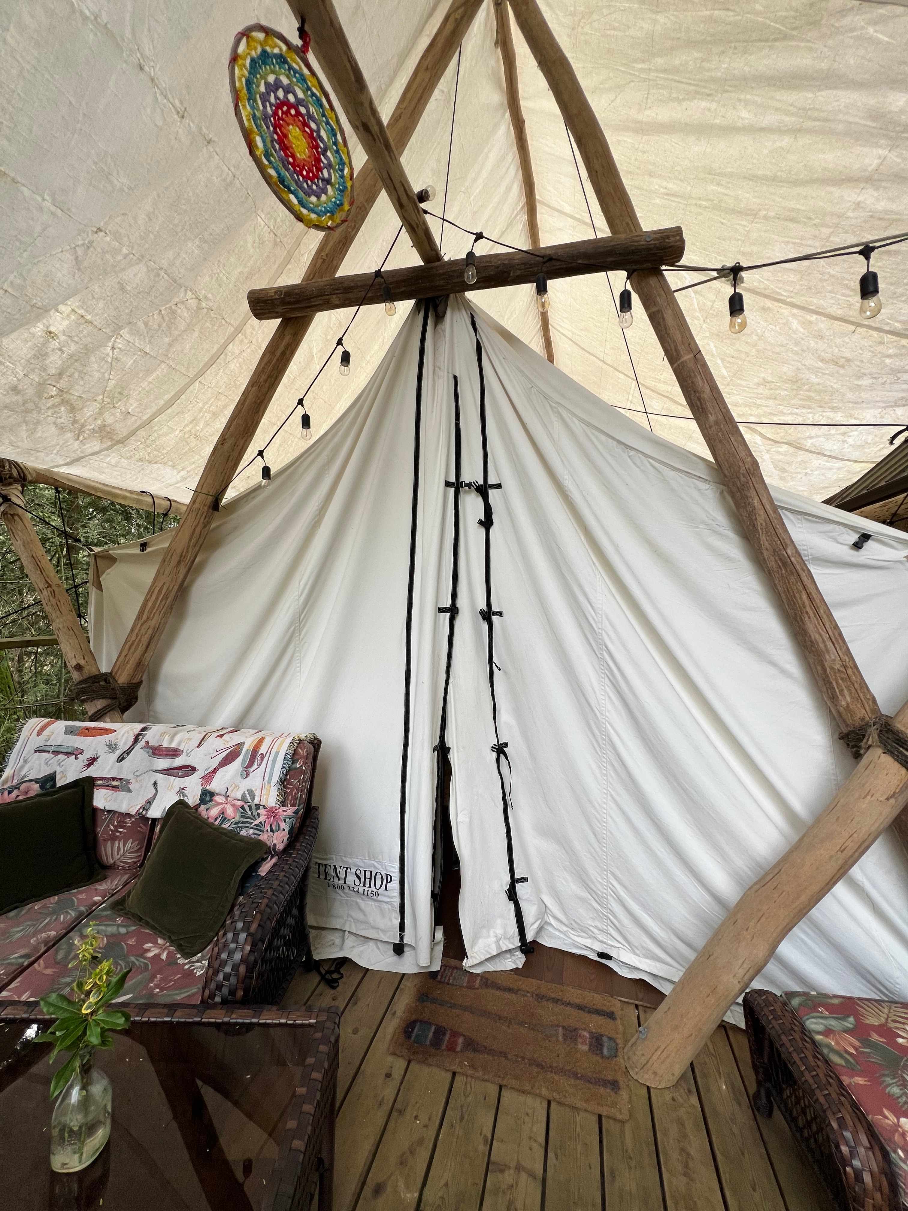 A white canvas tent pitched with bleached, smooth lumber, under a similar canopy. String lights hang decoratively above the tent wings, and a rainbow dreamcatcher hangs above the centre. A wicker loveseat set is placed in front of the tent, with matching table