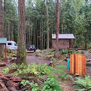 View of camping are with hytte in background, outdoor shower, outhouse, fire pit and picnic table.