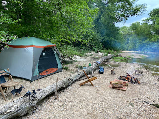 Green tent beside a gravel riverbank with a campfire, fallen log, and small dog nearby
