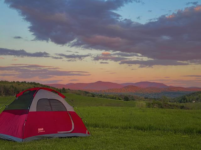 Red tent pitched in open field at dusk with mountains and colorful sky