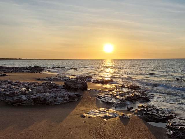 Sun setting over calm ocean, rocky shoreline silhouetted in warm golden light