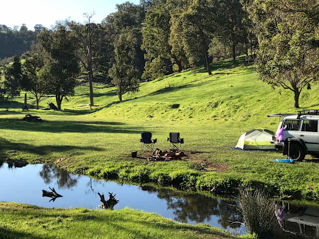 Two camp chairs and firepit beside a creek with tent and vehicle on green hillside at dawn