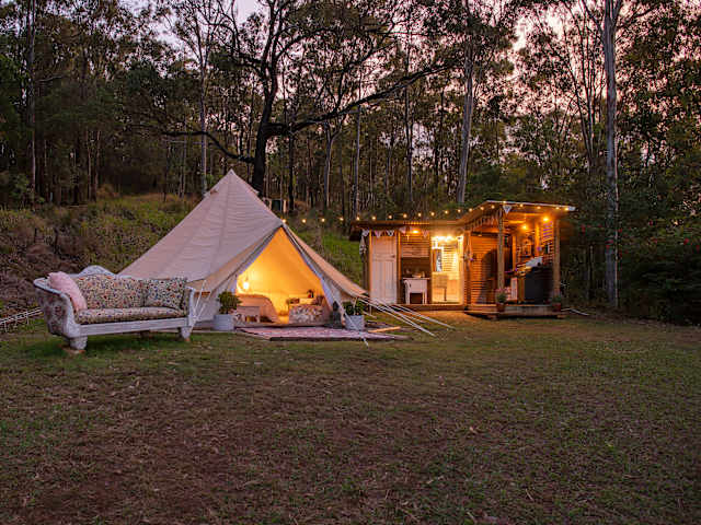 Bell tent and cabin lit up at dusk in bushland, outdoor sofa in foreground