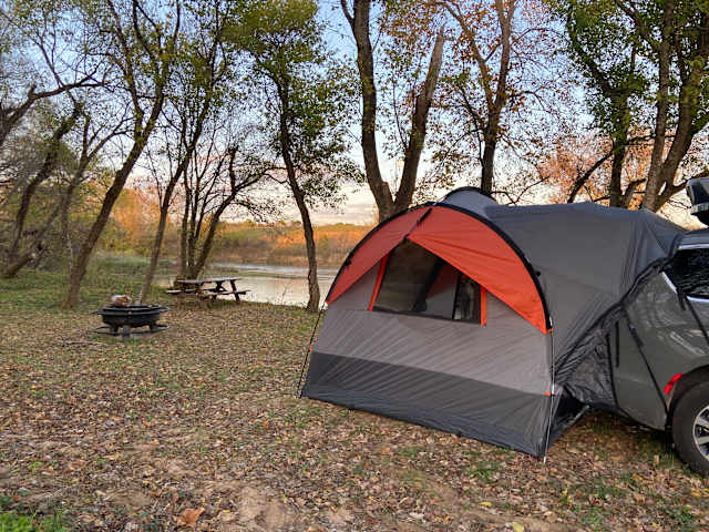 Orange and gray tent beside picnic table and fire ring, river visible through autumn trees at dusk
