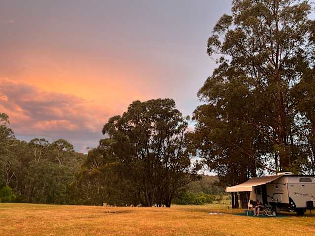 Caravan with awning at dusk under vivid orange and pink sunset clouds beside tall gum trees