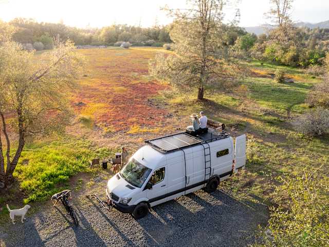 Aerial view of camper van with people on roof beside golden field of wildflowers at sunset