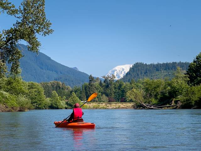 Person kayaking red kayak on calm river with forested mountains and snow-capped peak beyond