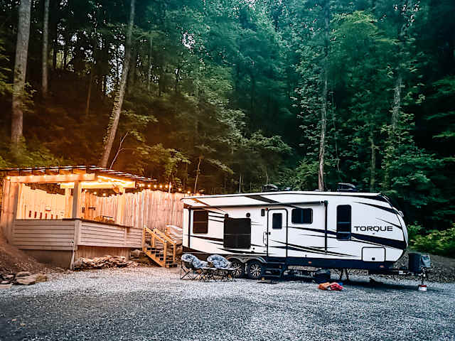 Travel trailer parked beside a wood-built outdoor kitchen strung with warm lights at dusk, forest behind