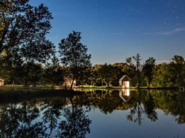 Cabin glowing at night beside a still pond under a starry sky, silhouetted trees