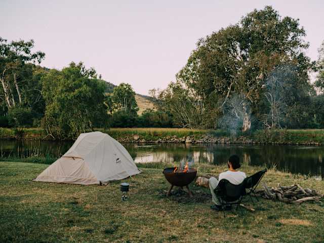 Tent and campfire beside a calm river at dusk, person seated by the flames, tree-lined hills beyond