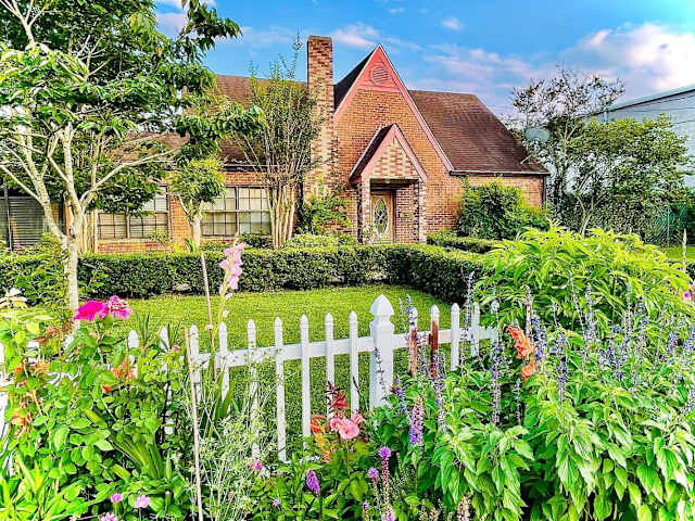 Brick cottage with white picket fence and colorful flower garden in foreground under blue sky