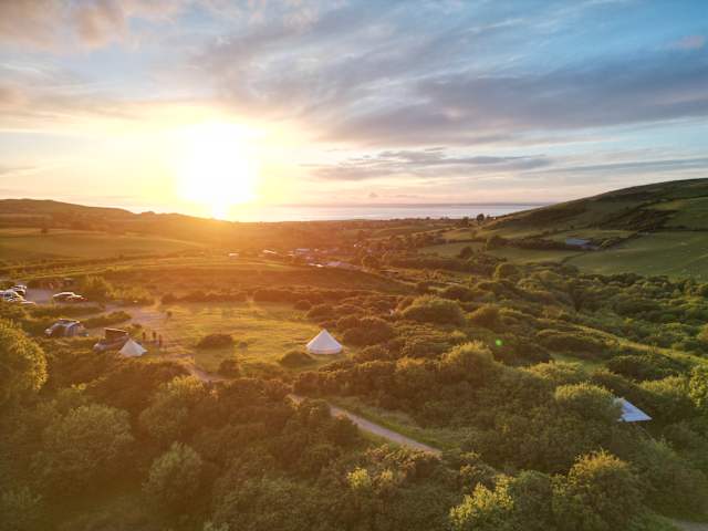 Aerial view at sunset of green Welsh hills, sea, and white bell tents nestled among hedgerows