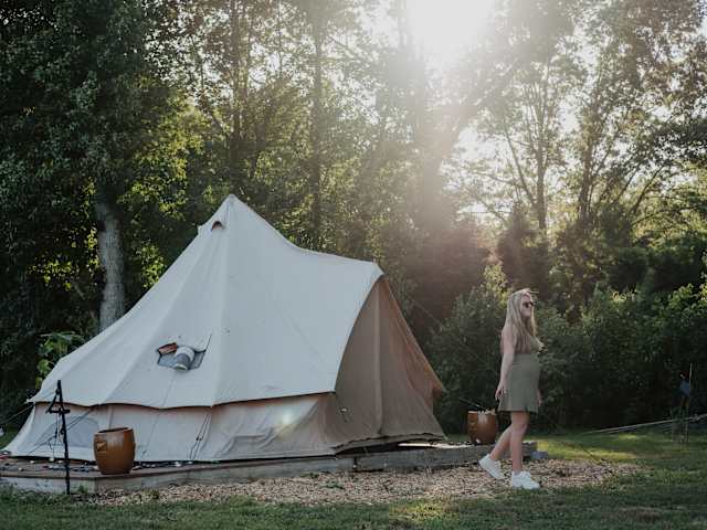 Woman walking beside a canvas bell tent lit by golden hour sunlight through trees