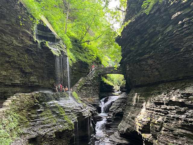 Layered rock gorge with waterfalls cascading beside a stone footbridge and green canopy