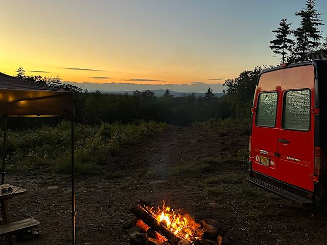 Red van parked beside campfire at dusk overlooking forested hills and sunset
