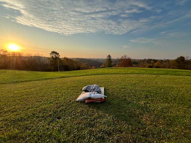 Pillows and blanket laid on grassy hilltop at sunset overlooking rolling countryside