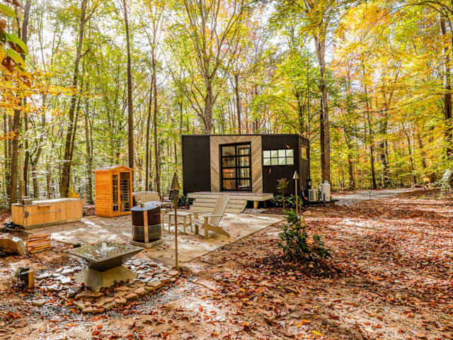 Black-and-wood tiny cabin in golden autumn forest with sauna, hot tub area, and fire pit on a stone patio