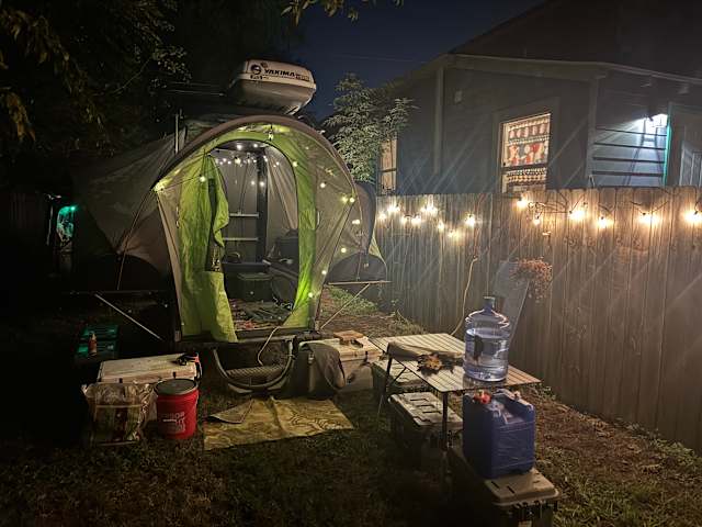 Green tent lit with string lights at dusk in a fenced backyard campsite