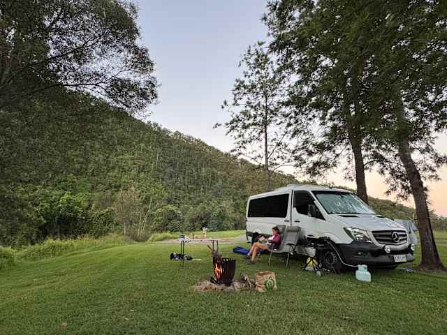 Campervan with awning parked at dusk beside unlit fire pit, forested hill behind, person reading in chair