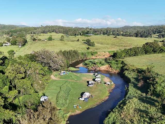 Aerial view of riverside campsite with vehicles and kayaks in golden light, river bend and hills beyond