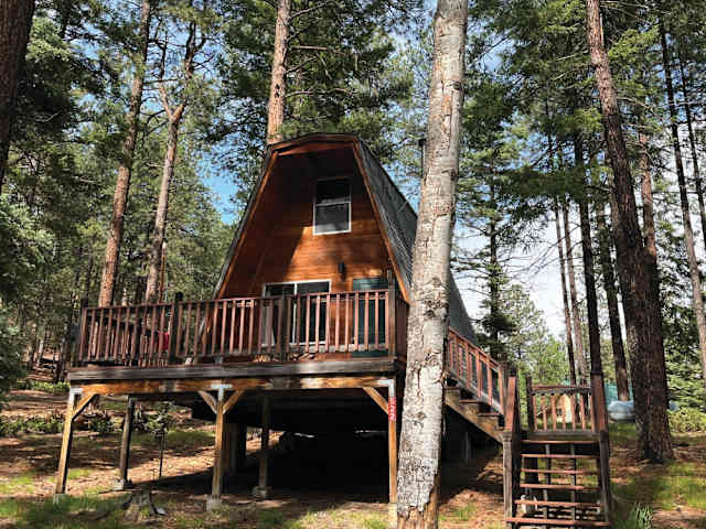 Wooden A-frame cabin on stilts surrounded by tall pine trees under blue sky