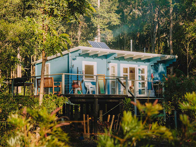 Blue timber cabin on stilts with solar panel and deck, surrounded by forest at golden hour