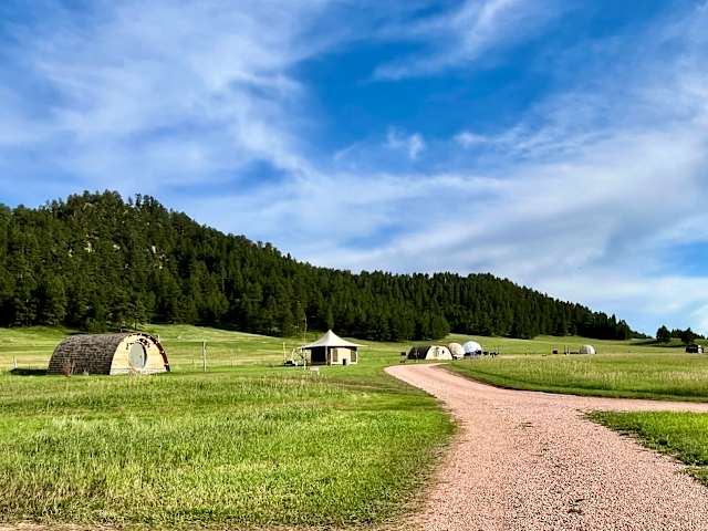 Gravel road curves through green meadow toward dome tents beneath pine-covered hills and blue sky