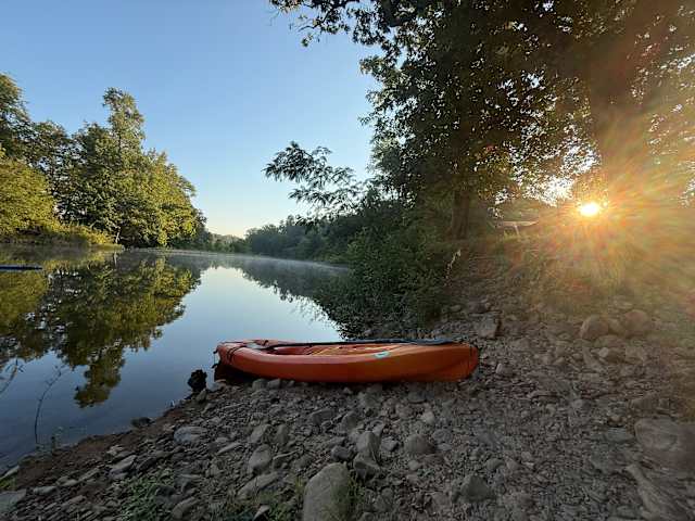 Orange kayak resting on rocky riverbank at sunrise with mist and glowing sun through trees