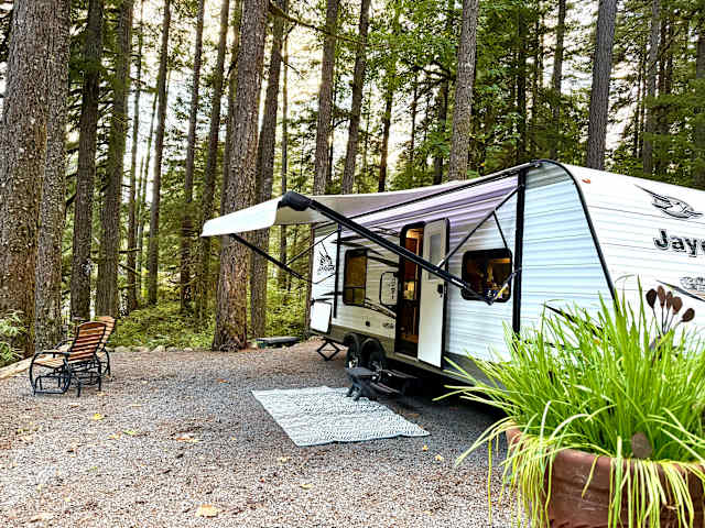 White travel trailer with awning open in forest campsite, chairs and rug outside