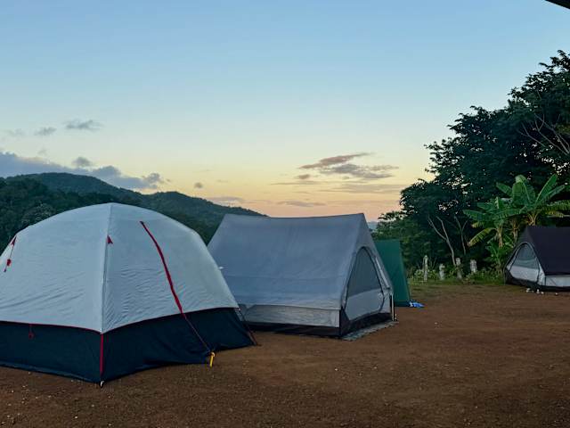 Three tents pitched on a ridge at dusk with mountains silhouetted against a soft sunset sky
