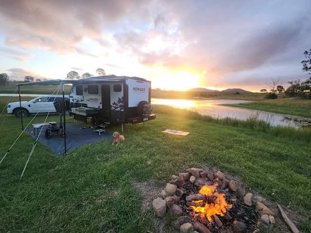 Camper trailer and campfire beside a lagoon at golden sunset with a dog resting nearby