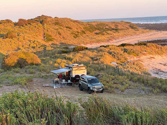 Camper trailer with awning on grassy dune at sunset, ocean and beach beyond, warm light