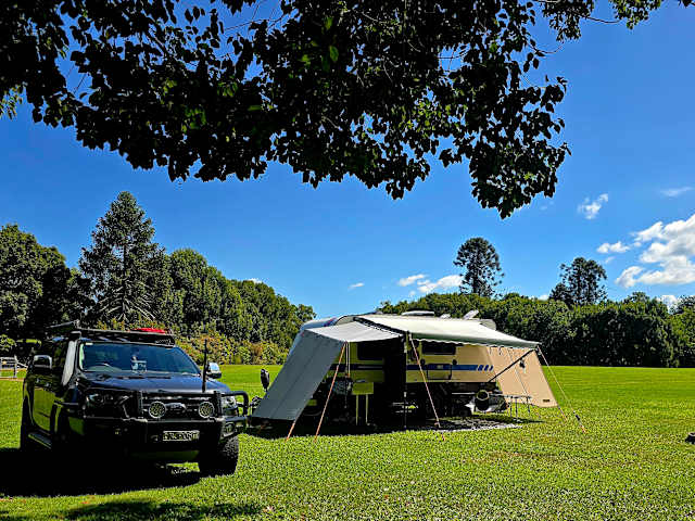 Camper trailer with awning and 4WD parked under leafy tree canopy on green grass under blue sky