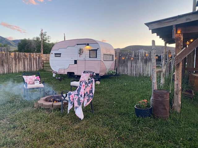 Pink vintage camper at dusk with lit fire pit, chairs, and mountain backdrop