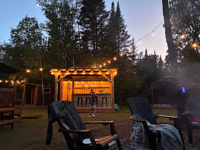 Wooden pavilion bar lit with string lights at dusk, person dancing, forest backdrop