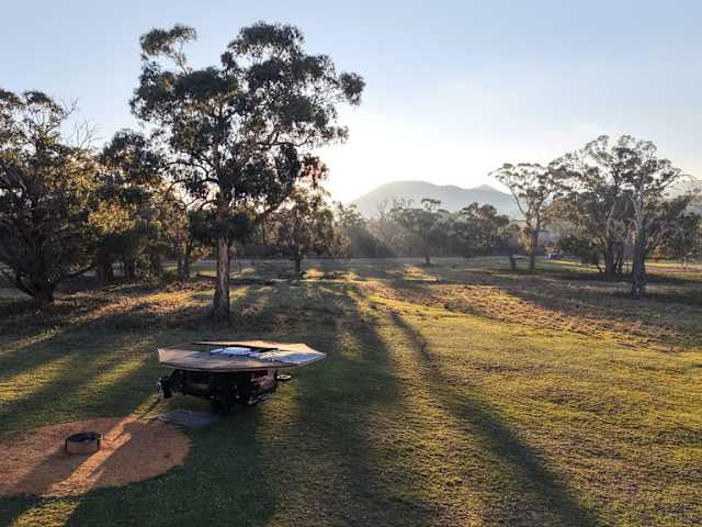 Rooftop-tent trailer parked in grassy paddock at sunrise with mountain and gum trees