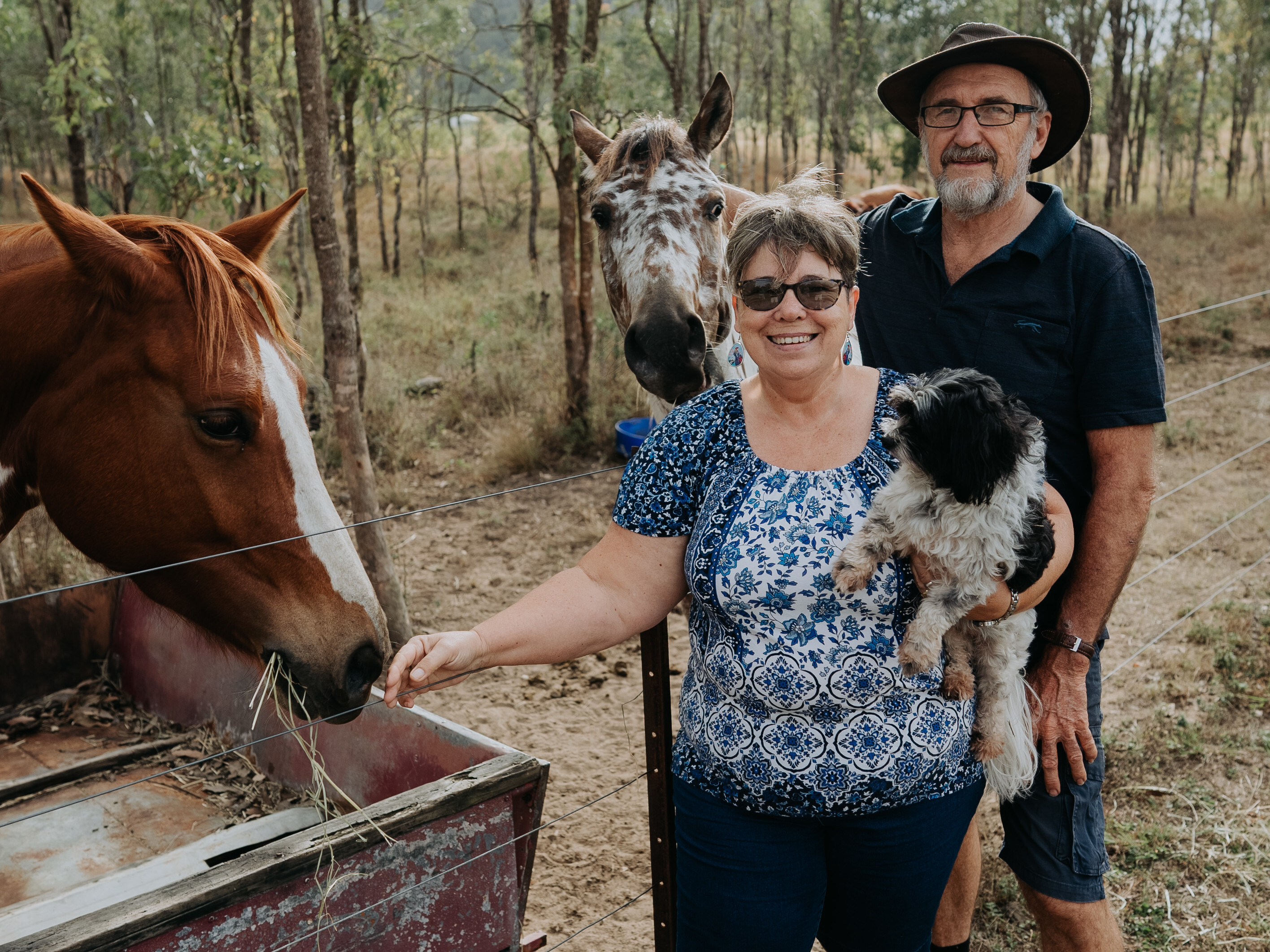A woman is standing in front of her husband. The woman is holding a small black and white dog and is patting a chestnut horse. Behind them is another horse.
