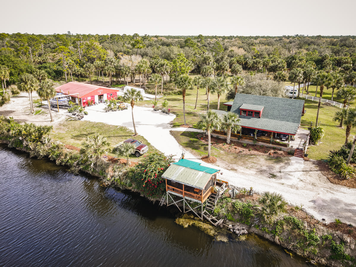 The property borders the beautiful river. You can see the screened open-air kitchen alongside the water, the main house, and the red greenhouse 