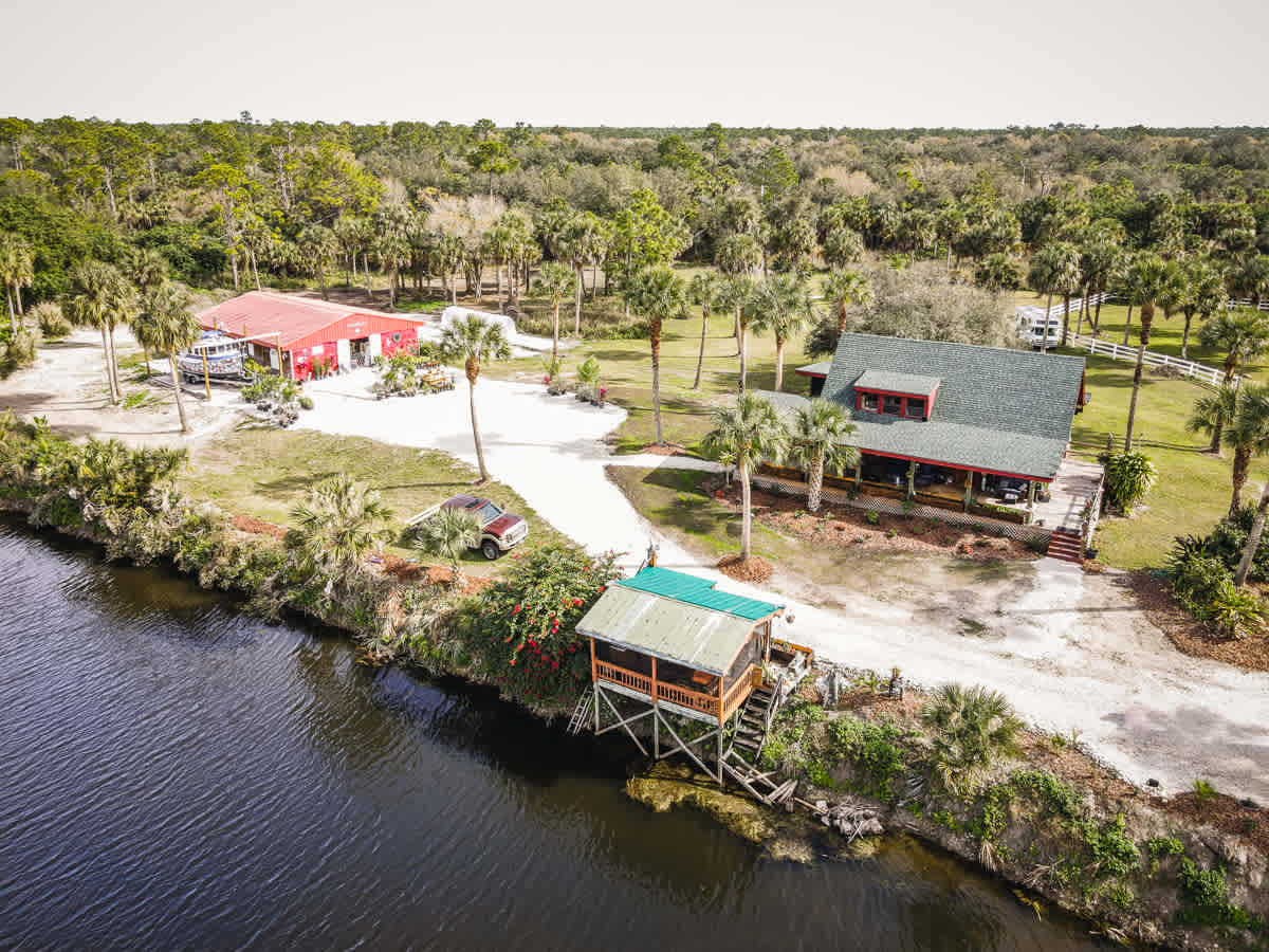The property borders the beautiful river. You can see the screened open-air kitchen alongside the water, the main house, and the red greenhouse 